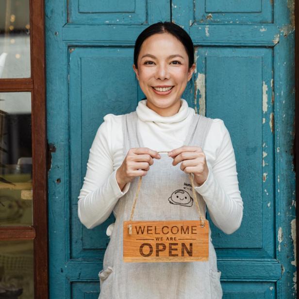Eine lächelnde Frau hält ein Schild mit der Aufschrift „Welcome – We are Open“ vor einer blauen Tür.