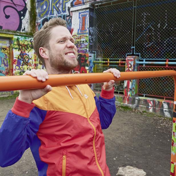 Ein Mann in einer bunten Jacke macht Klimmzüge an einer orangefarbenen Stange auf einem Spielplatz.