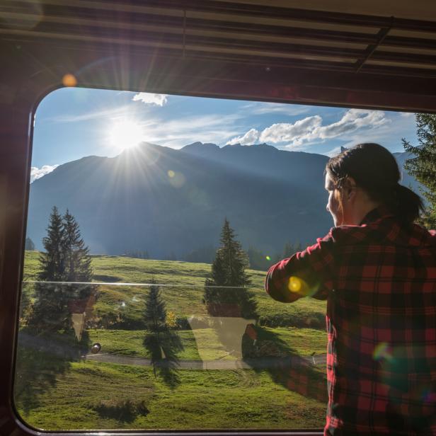 Eine Frau blickt aus dem Fenster eines Zuges auf eine grüne Berglandschaft.