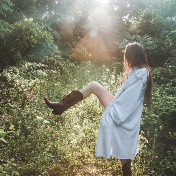 Eine Frau mit Morgenmantel und Cowboystiefeln steht in der Natur.