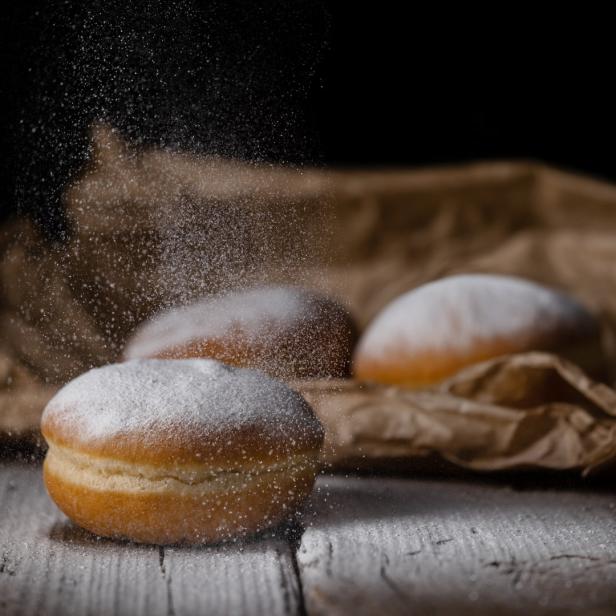 Drei Krapfen liegen auf einem Holztisch, während Puderzucker darüber gestreut wird.