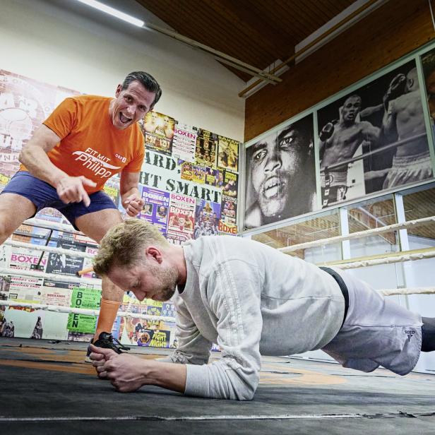 Ein Mann macht Plank-Übungen in einem Boxring, während ein Trainer ihn anfeuert.