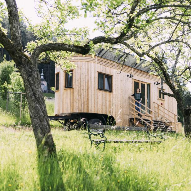 Eine Frau steht vor einem Tiny House auf Rädern in einer grünen Landschaft.