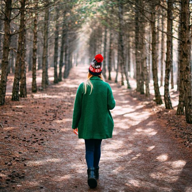 Eine Frau geht in einem grünen Mantel und einer Mütze mit Bommel durch einen Wald.