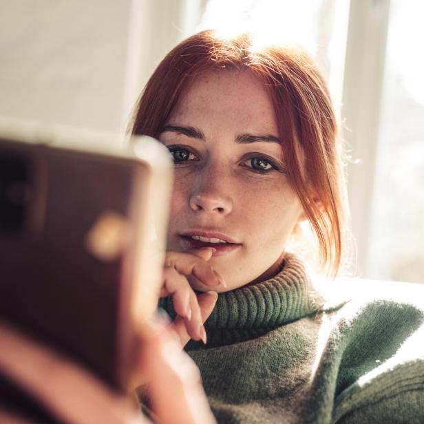 Frau mit roten Haaren schaut auf den Bildschirm ihres Mobiltelefons.