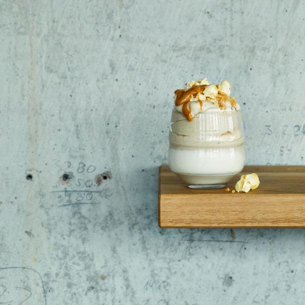 Ein Glas Dessert mit Karamell, Popcorn und einer hellen Creme steht auf einem Holzbrett.