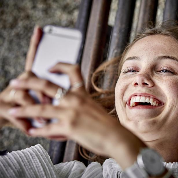 Eine lachende junge Frau liegt auf einer Bank und benutzt ihr Handy.