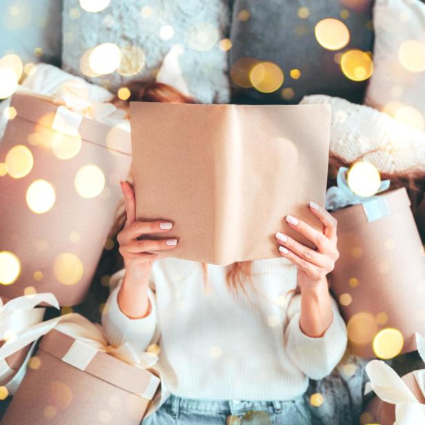 Eine schöne Frau liest ein Buch und liegt zwischen Geschenkboxen, während sie Weihnachtsangebote genießt.