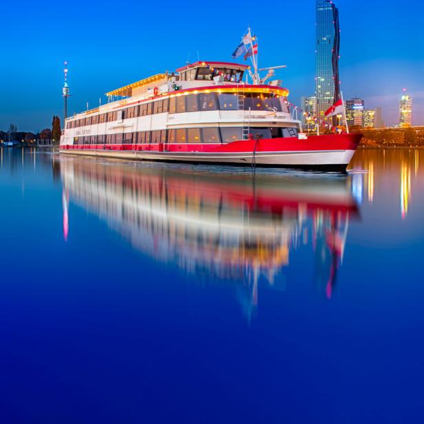 Ein Ausflugsschiff auf der Donau in Wien vor der Skyline der Stadt.