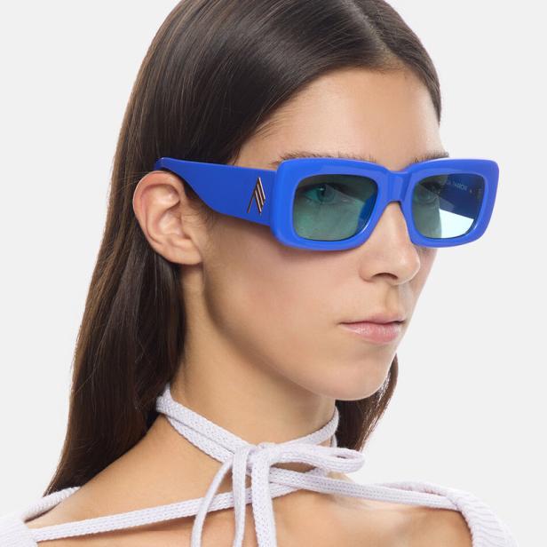 Eine Frau mit blauen, rechteckigen Sonnenbrillen und einem weißen Oberteil mit Schnürung.