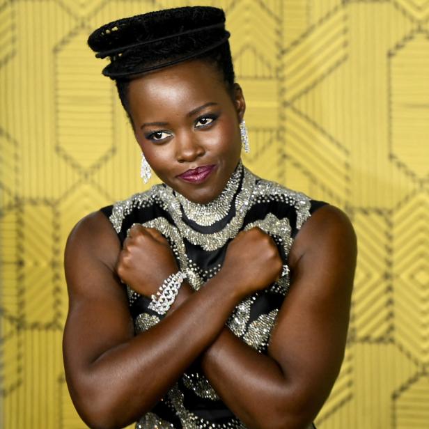 Lupita Nyong'o kreuzt die Arme vor der Brust, vor einem gelben Hintergrund.