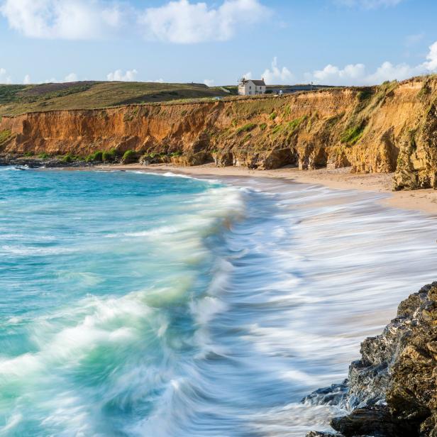 Türkisfarbenes Wasser umspült einen Sandstrand mit Klippen im Hintergrund.