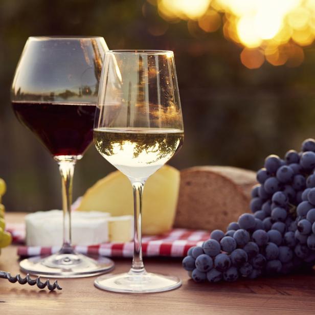 Zwei Gläser Wein, Käse, Brot und Trauben auf einem Tisch im Freien.