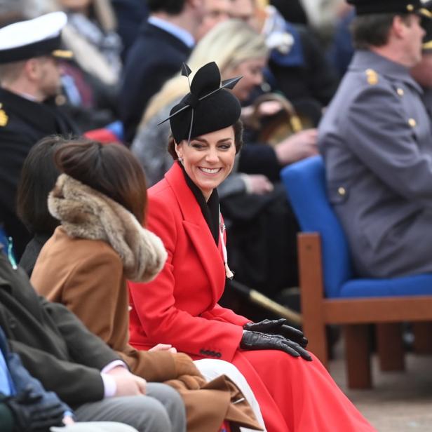 Kate Middleton sitzt in einem roten Mantel und einem schwarzen Hut lächelnd in der Menge.