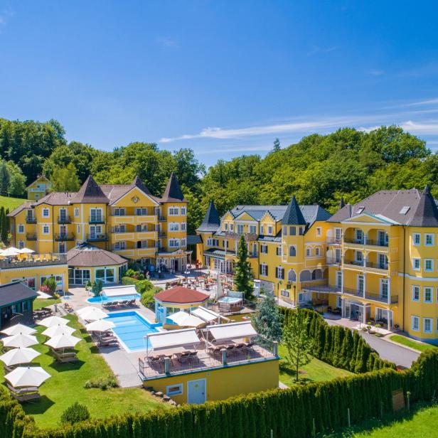 Ein gelbes Hotelresort mit Pool und gepflegten Gärten vor einem bewaldeten Hügel.