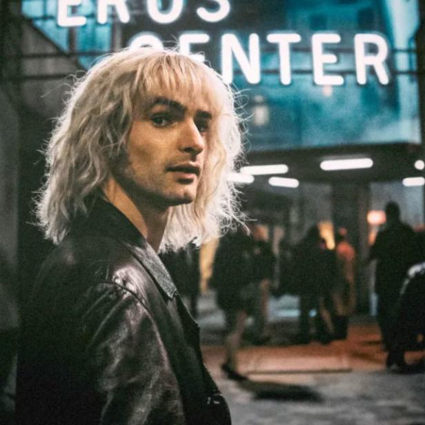 Ein Mann mit blonden Haaren steht vor einem Gebäude mit der Aufschrift „Eros Center“.