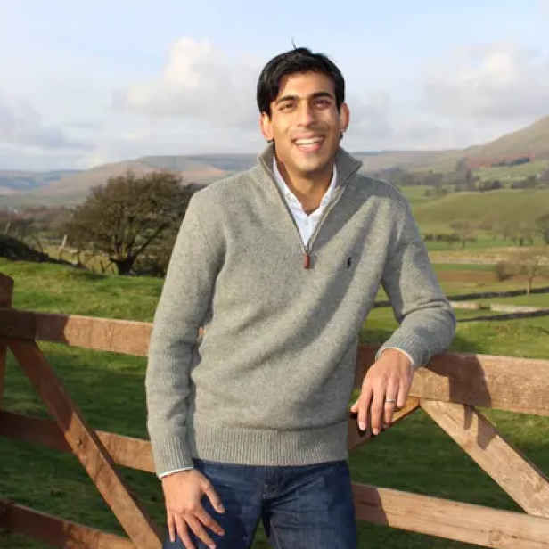 Rishi Sunak lächelt vor einer ländlichen Kulisse in die Kamera.