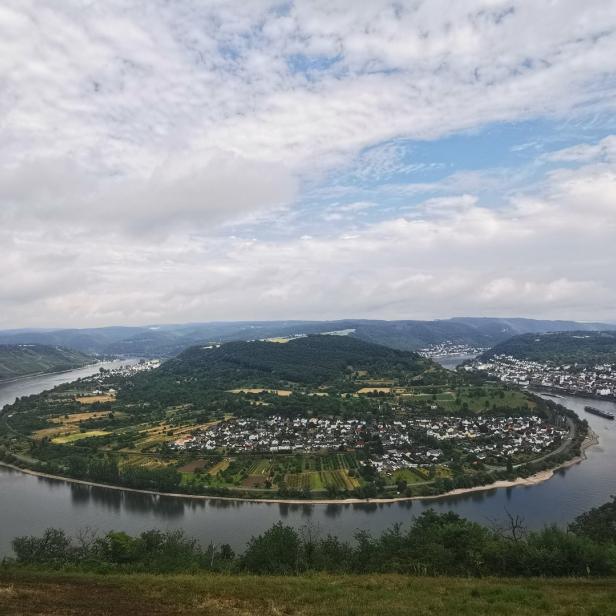 Blick auf eine Flussschleife mit einer Ortschaft und bewaldeten Hügeln unter bewölktem Himmel.