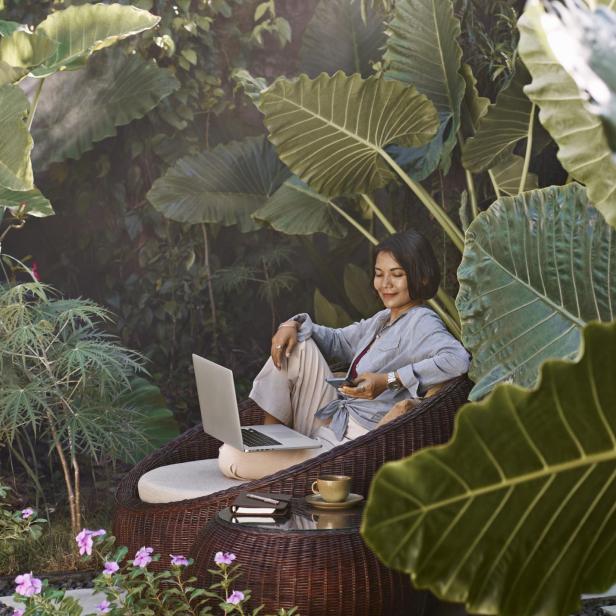 Eine asiatische Frau arbeitet von zu Hause aus, sitzt in Gartenmöbeln, umgeben von tropischen Pflanzen, und benutzt einen Laptop und ein Mobiltelefon.