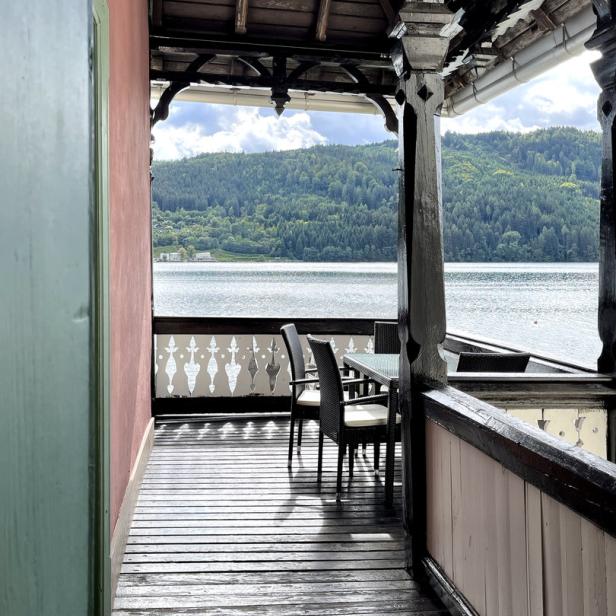 Blick von einer hölzernen Veranda auf einen See und bewaldete Hügel.