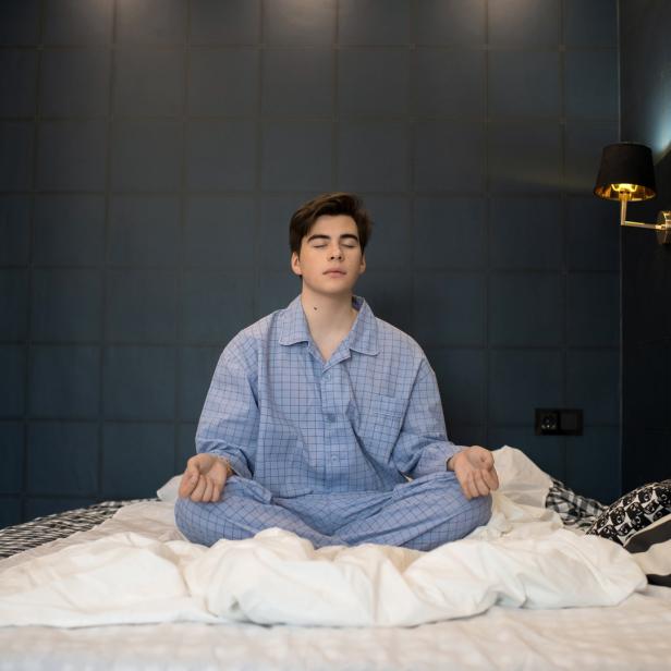 Ein Mann im Pyjama sitzt im Schneidersitz auf einem Bett und meditiert.