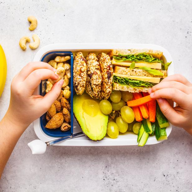 Eine geöffnete Jausenbox mit Sandwich, Obst, Gemüse und Nüssen wird von Kinderhänden befüllt.