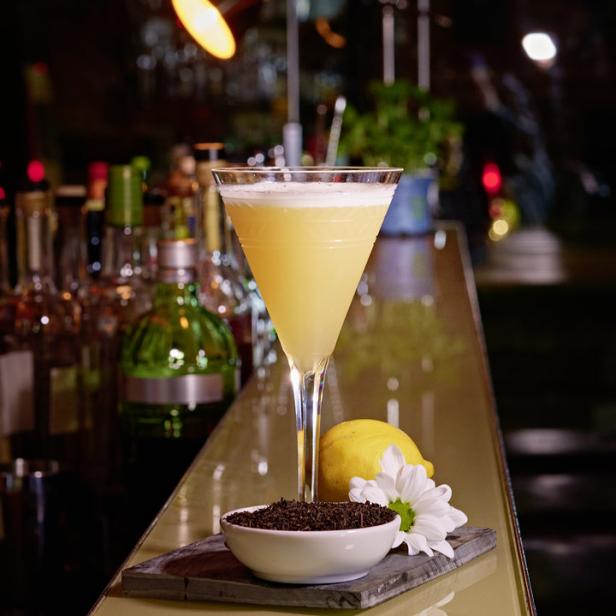 Ein Cocktail in einem Martiniglas steht auf einer Bar, garniert mit Zitrone, Blüte und einer Schale Tee.