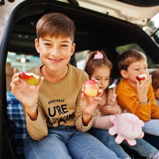 Familie mit vier Kindern isst Äpfel im Fahrzeuginnenraum. Kinder sitzen im Kofferraum. Anreise mit dem Auto in die Berge, Atmosphärenkonzept.