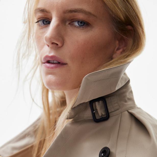 Eine blonde Frau trägt einen beigefarbenen Trenchcoat mit hohem Kragen.