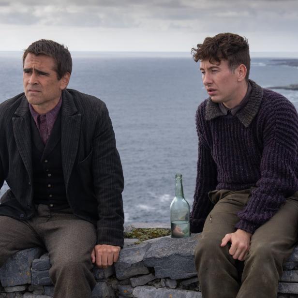 Colin Farrell und Barry Keoghan sitzen mit einer Flasche auf einer Steinmauer vor einer Küstenlandschaft.