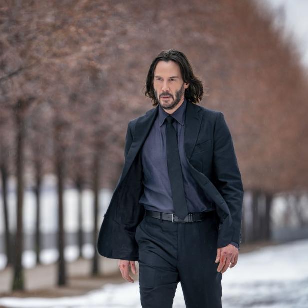 Keanu Reeves in einem dunklen Anzug geht auf einem verschneiten Weg entlang einer Baumreihe.