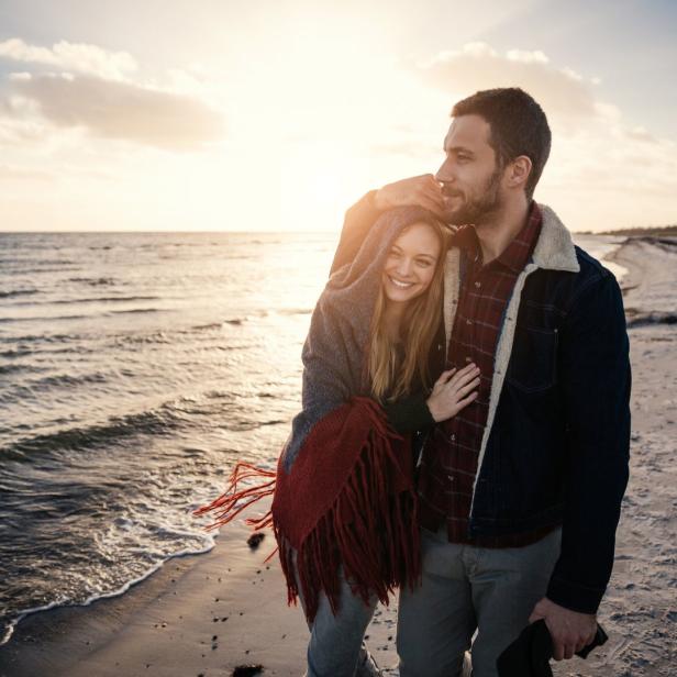 Ein Paar steht am Strand, in eine Decke gehüllt, während die Sonne untergeht.