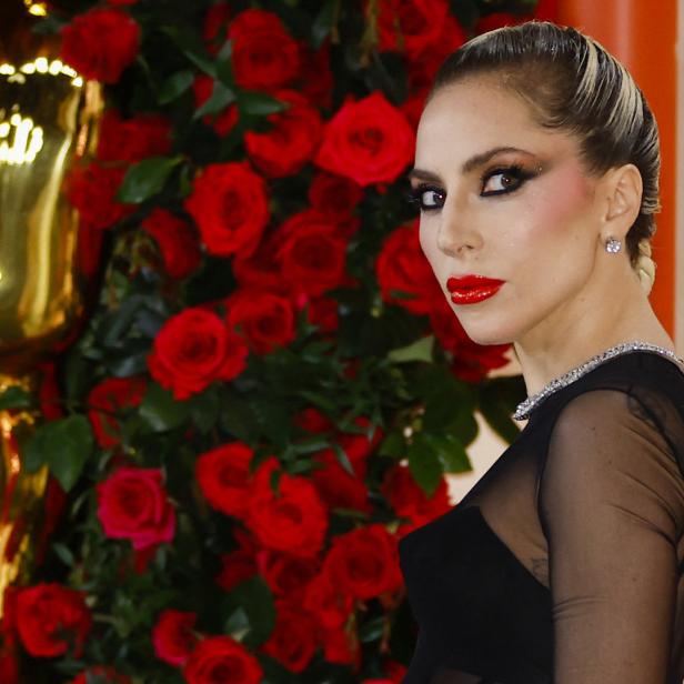 Lady Gaga posiert vor einer Wand aus roten Rosen und einer goldenen Statue.