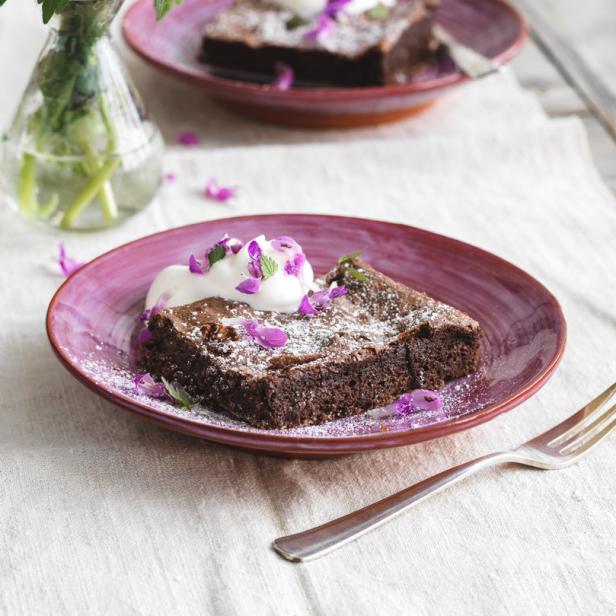 Ein Schokoladenbrownie mit Puderzucker, Sahne und lila Blüten auf einem Teller.