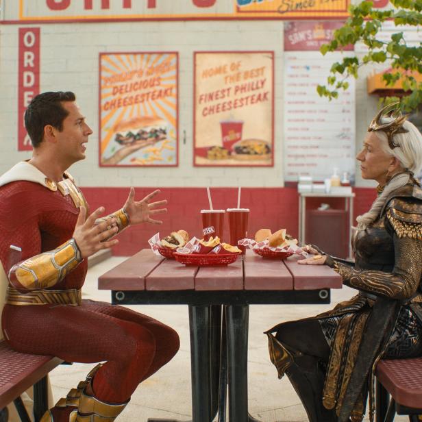 Shazam und Hespera sitzen an einem Tisch vor einem Imbiss und essen Cheesesteaks.