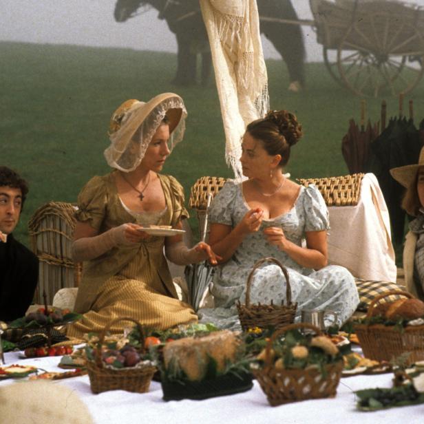 Eine Picknickgesellschaft in Kostümen des frühen 19. Jahrhunderts sitzt auf einer Wiese.
