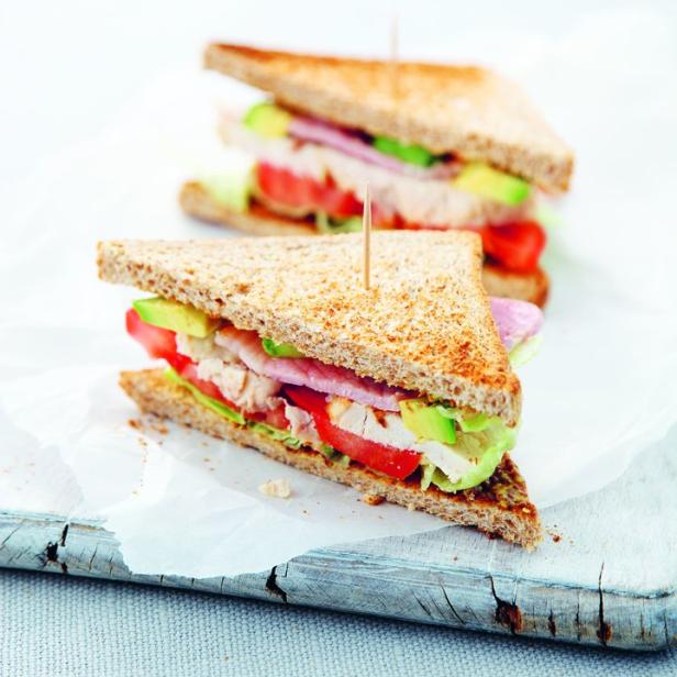 Zwei angeschnittene Sandwiches mit Hähnchen, Avocado, Tomaten und Salat auf einem Holzbrett.
