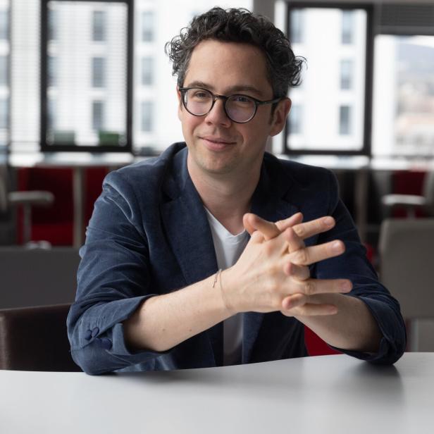 Ein Mann mit Brille und lockigen Haaren sitzt lächelnd an einem Tisch in einem modernen Büro.
