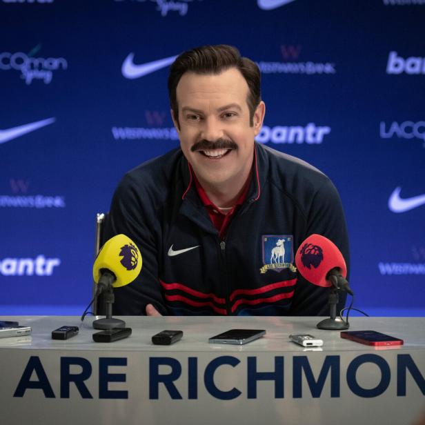 Ted Lasso sitzt lächelnd bei einer Pressekonferenz von AFC Richmond.