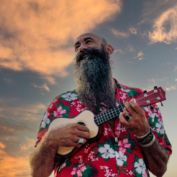 Rasierter Mann mit langem Bart spielt Ukulele vor einem dramatischen Himmel.