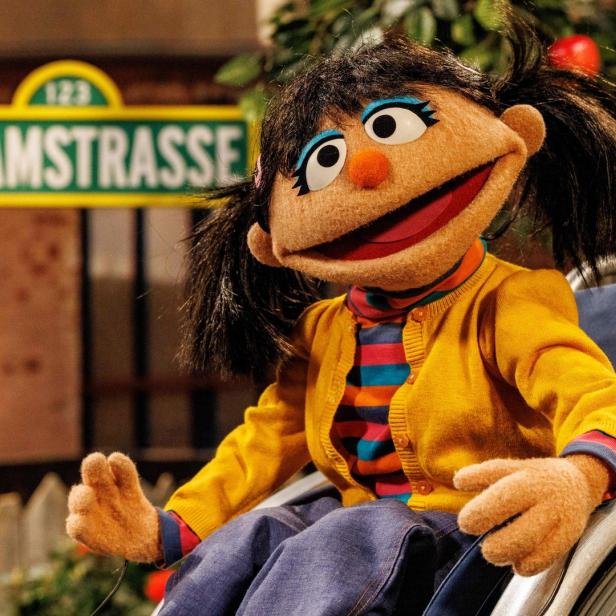 Eine Puppe der „Sesamstraße“ sitzt in einem Rollstuhl vor dem Straßenschild.