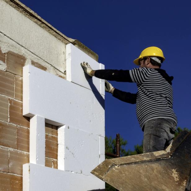 Ein Bauarbeiter mit gelbem Helm befestigt Dämmplatten an einer Hauswand.