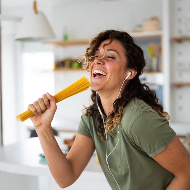 Eine Frau singt mit einem Bund Spaghetti in der Hand in ihrer Küche.