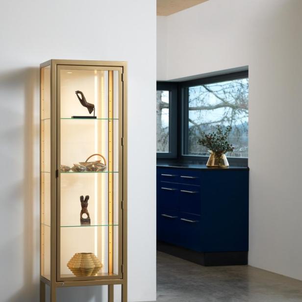 Ein goldfarbener Vitrinenschrank mit Glasböden und verschiedenen Dekorationsgegenständen steht in einem Raum.