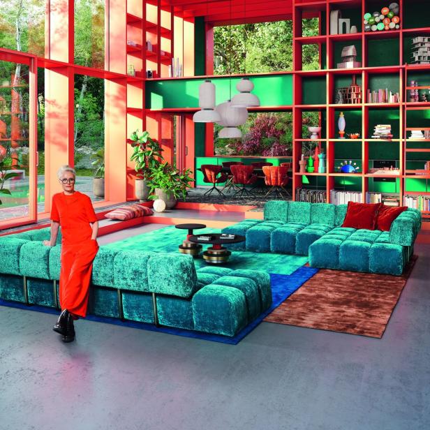 Eine Frau in einem orangefarbenen Overall lehnt an einem türkisfarbenen Sofa in einem farbenfrohen Wohnzimmer.