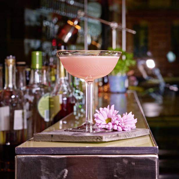 Ein rosa Cocktail in einem Glas mit Stiel, garniert mit einer rosa Blume auf einer Bar.