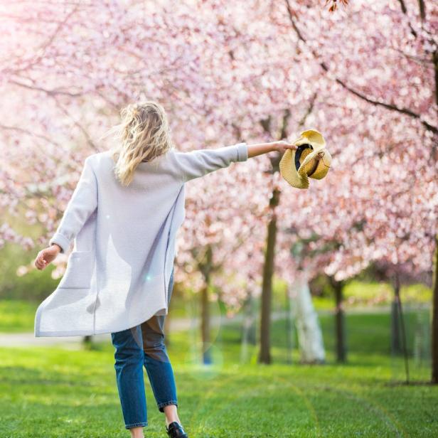 Eine Frau geht mit einem Hut in der Hand durch einen Park mit blühenden Bäumen.