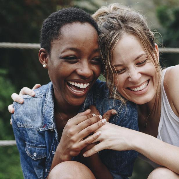 Zwei lachende Frauen umarmen sich vor einem grünen Hintergrund.