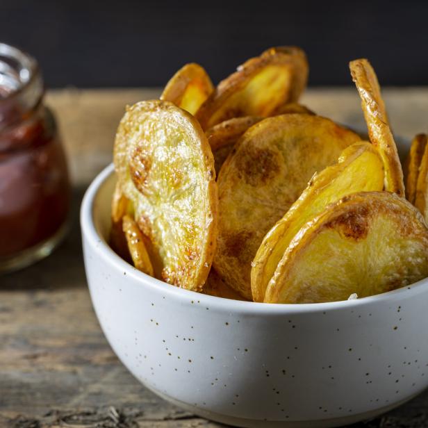 Eine Schüssel mit hausgemachten Kartoffelchips und ein Glas Ketchup stehen auf einem Holztisch.