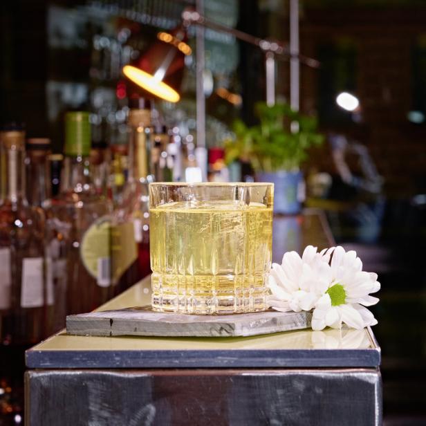 Ein Cocktail in einem Kristallglas steht neben einer weißen Blüte auf einer Bar.